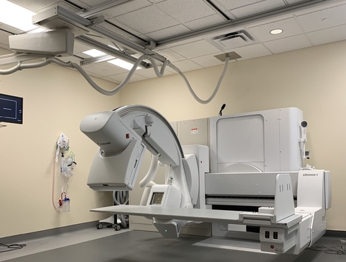 A fluoroscopy machine in a hospital imaging room. The large, C-shaped arm is positioned over a flat patient table, with overhead cables and monitors visible. The equipment is designed for real-time X-ray imaging to assist with diagnostic and interventional procedures. The room is clean and well-lit, with minimal furnishings.